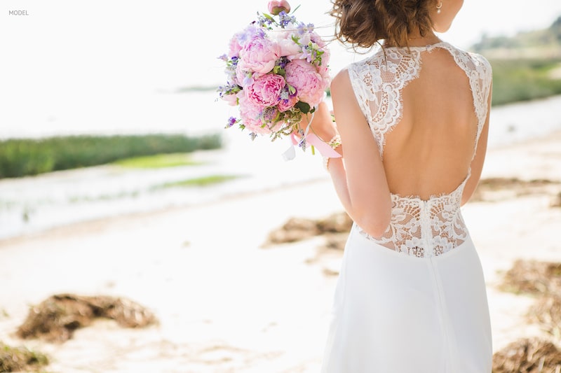 Image of bride's back with a lacy dress, standing on a rocky beach.
